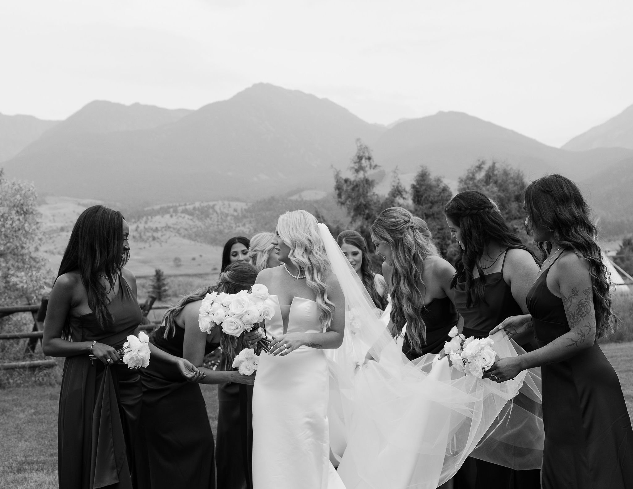 The bride, holding a bouquet of white flowers, stands surrounded by her bridesmaids in front of the stunning Montana mountains at Copper Rose Ranch Wedding Venue. The photo captures the natural beauty and emotional atmosphere of the wedding day, taken by Haley J Photo, a Montana Wedding Photographer.