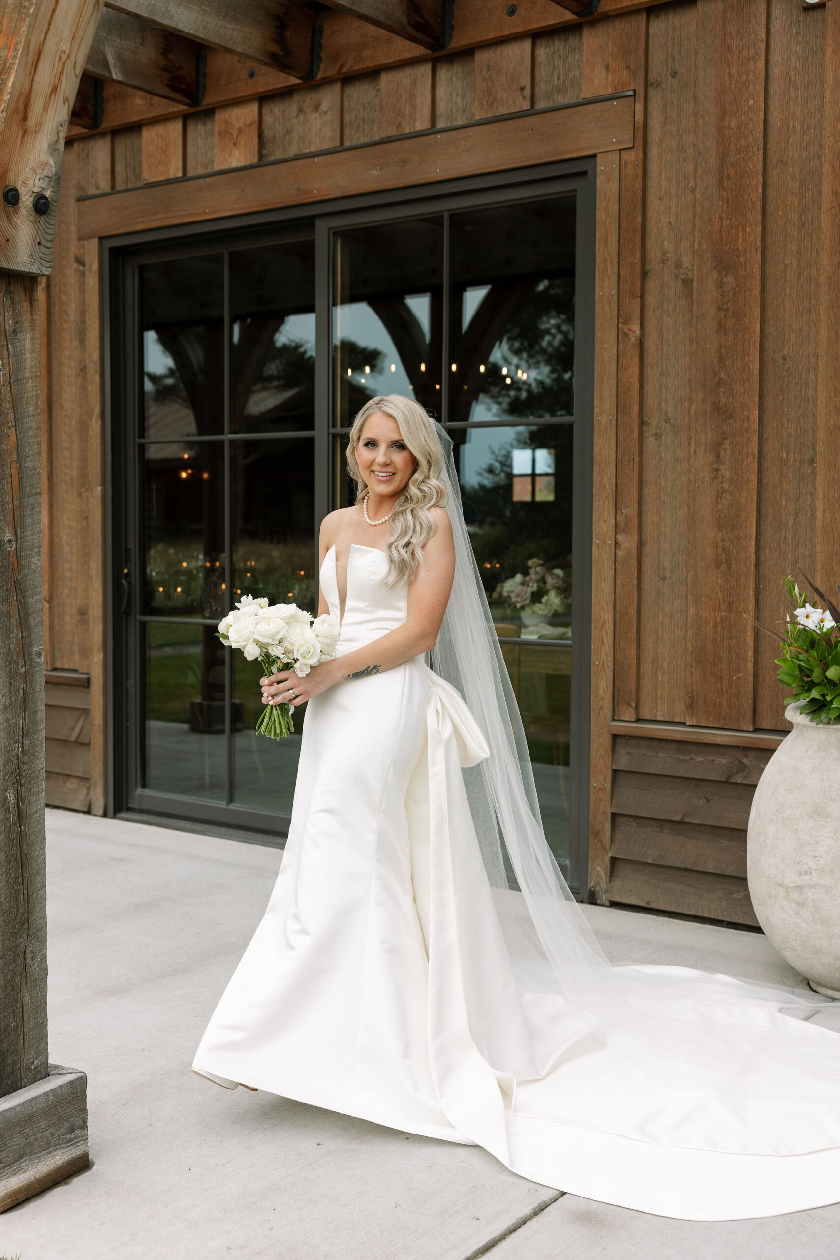 A bride in a white, strapless gown and long veil, holding a bouquet of white flowers. She stands on the porch of the rustic venue, smiling softly and looking serene.