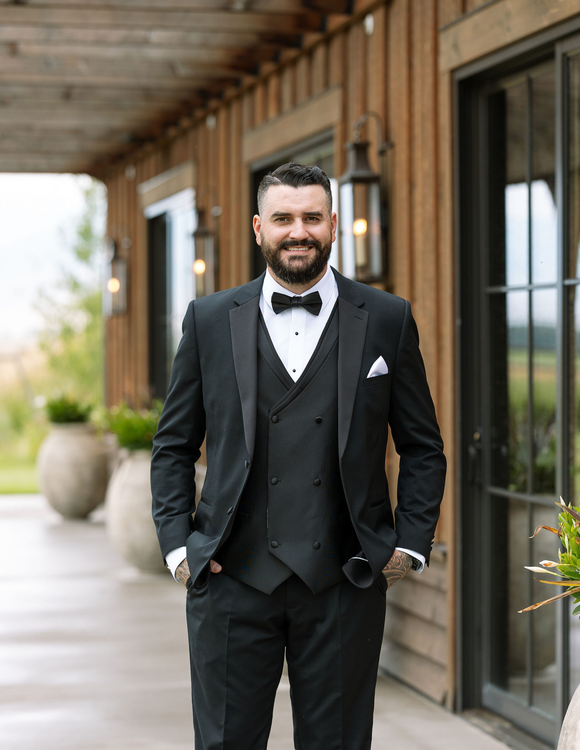 A groom smiling confidently, dressed in a classic black tuxedo with a white pocket square, standing on a wooden porch with the venue's rustic exterior in the background.