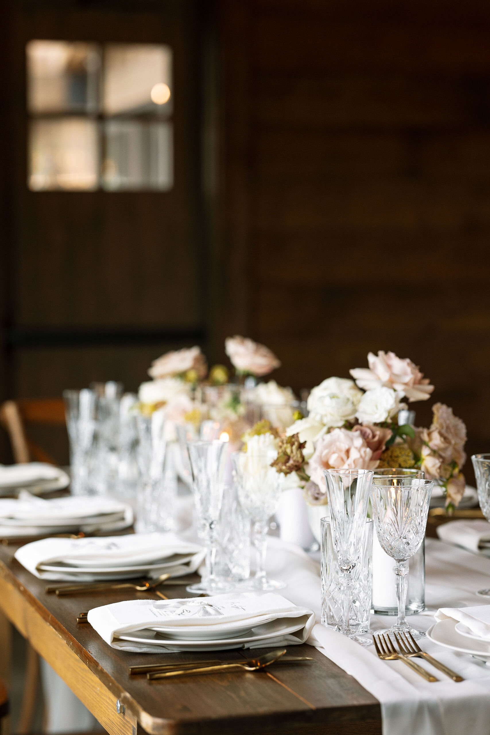  beautifully set dining table with crystal glassware, white and blush floral centerpieces, and elegant place settings, ready for a wedding reception. The warm wooden tones of the venue enhance the ambiance.