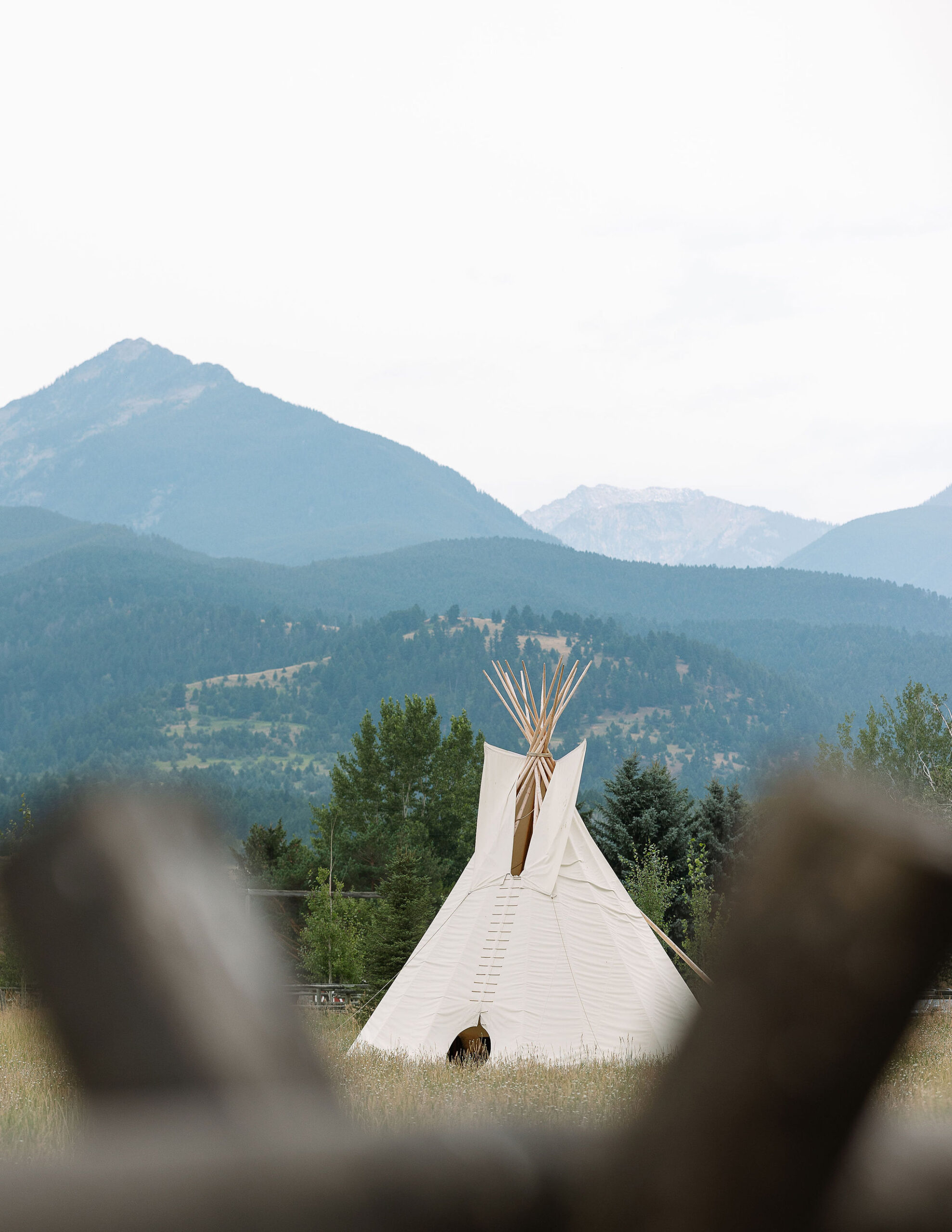 A scenic Montana Wedding Venue view of a tall, white teepee set against a backdrop of rolling hills and towering mountains under a cloudy sky.