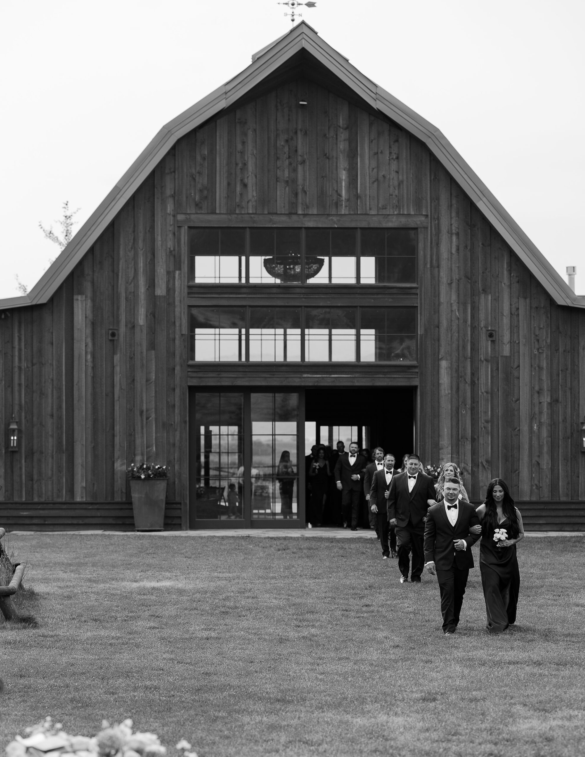 A black and white photograph showing a line of groomsmen walking towards a large wooden barn with a tall, peaked roof. The mood is formal and reflective.