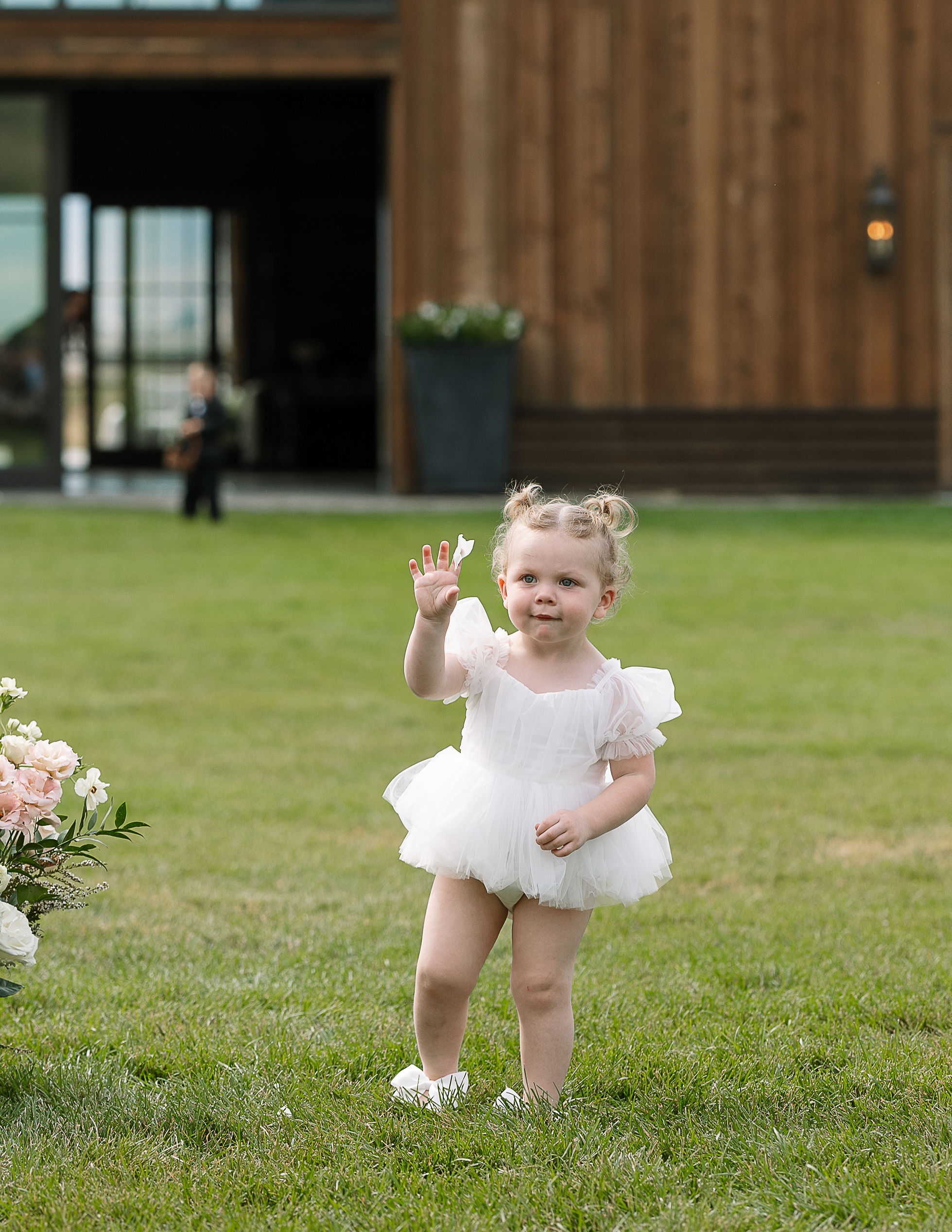  A young girl, dressed in a white ruffled dress, waves while standing on a lawn. The background includes a rustic wooden building.