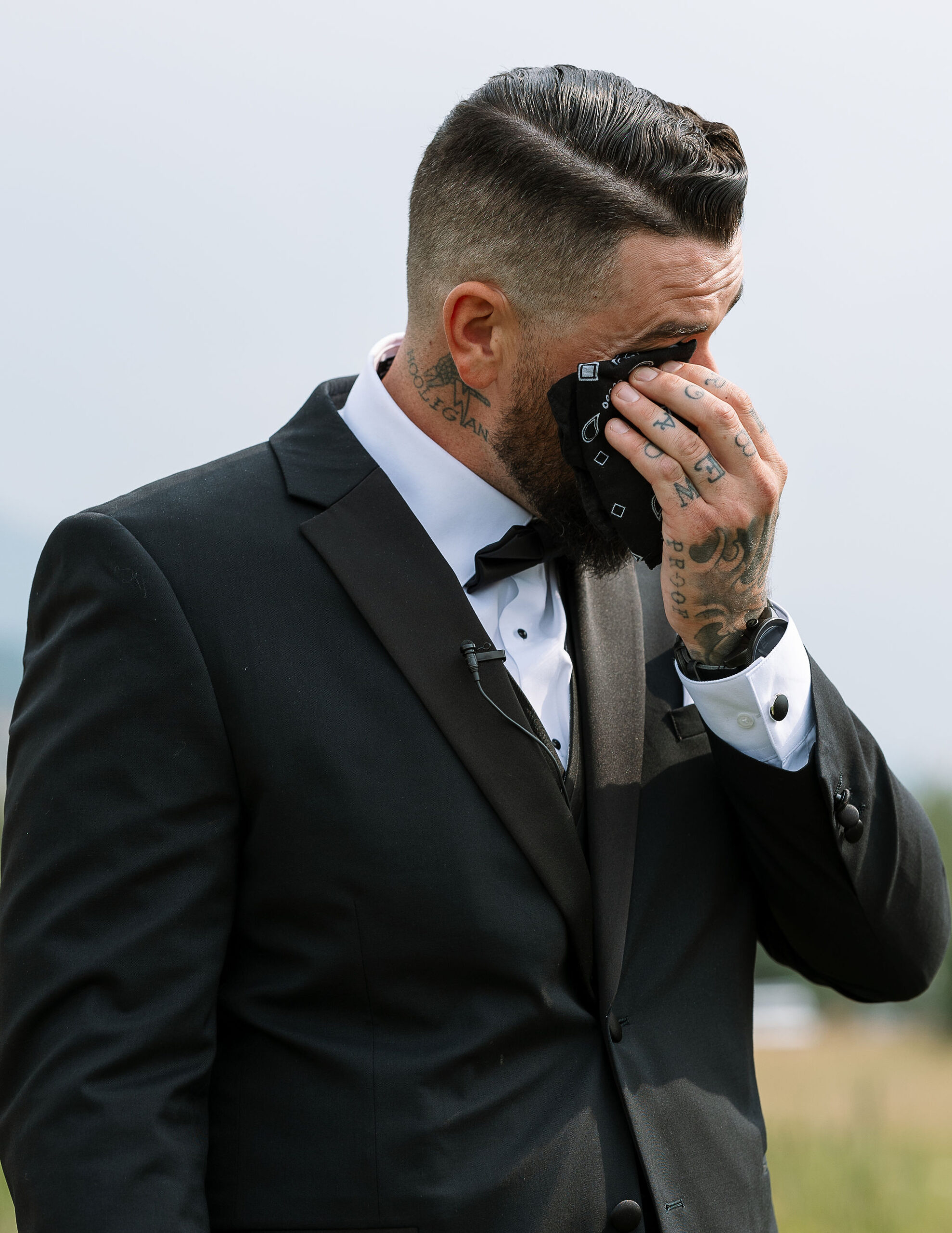 A groom in a black tuxedo and white shirt, overcome with emotion, wipes a tear from his eye with a black handkerchief as he stands outside in a natural setting.