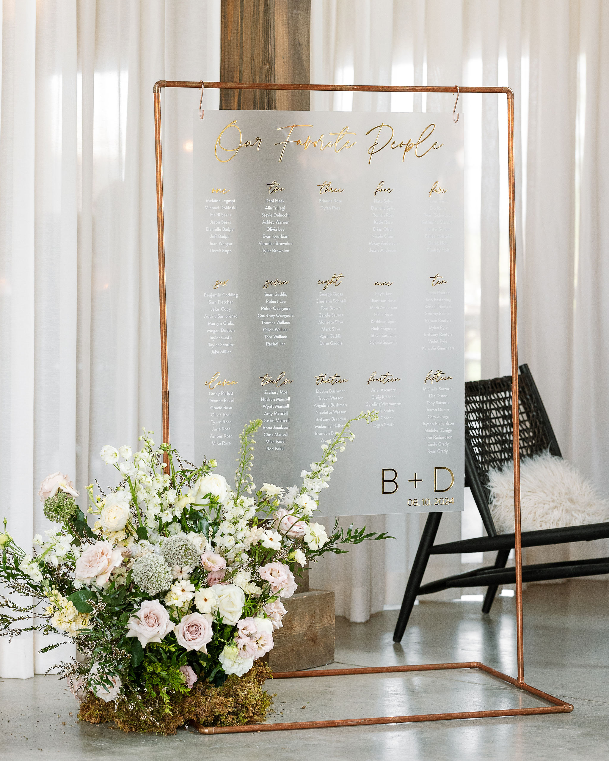 A seating chart display made of clear acrylic with gold lettering, set in a copper frame. A lush floral arrangement in soft pink and white hues sits at the base, while a minimalist black chair is visible in the background.