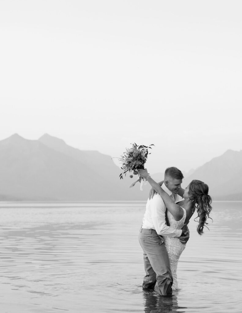 Bride and groom embracing in the water, with mountains in the background, at their Glacier National Park elopement.