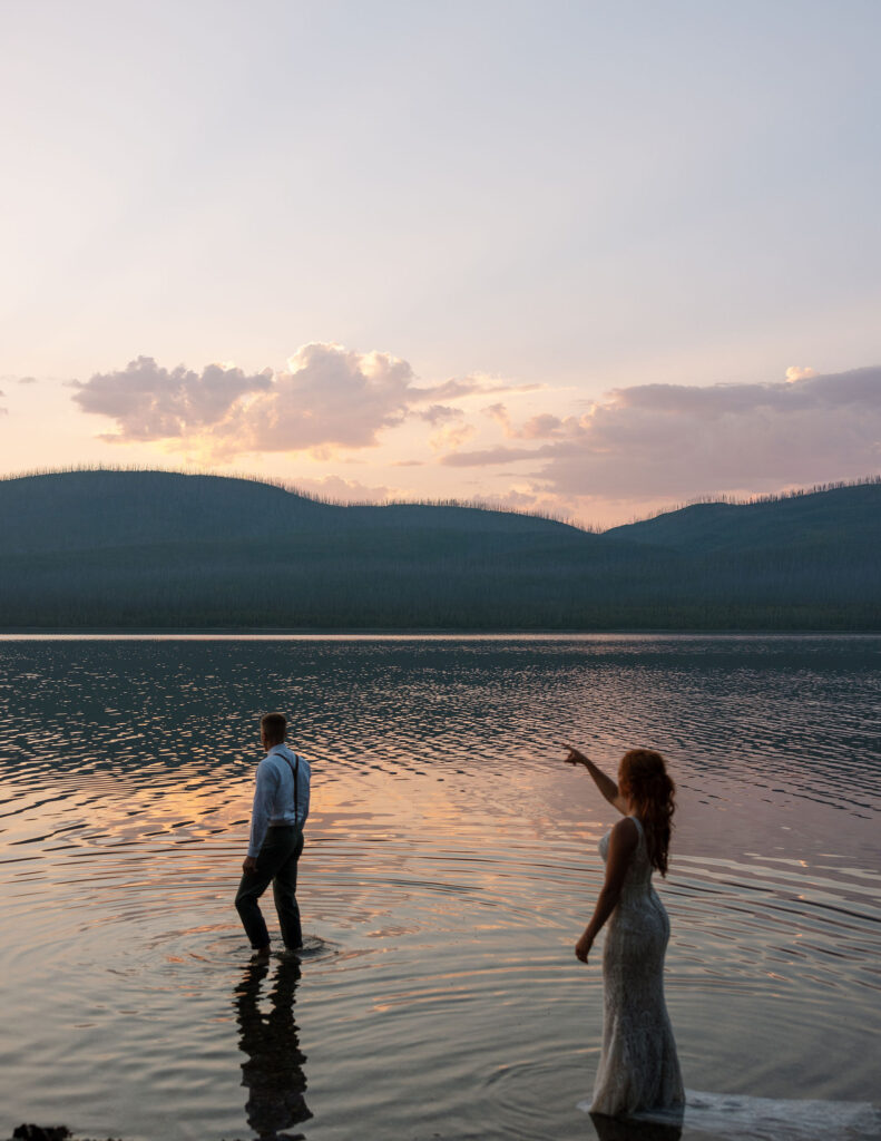 Bride and groom wading in the water at dusk, with scenic mountains in the background, during their Glacier National Park elopement.