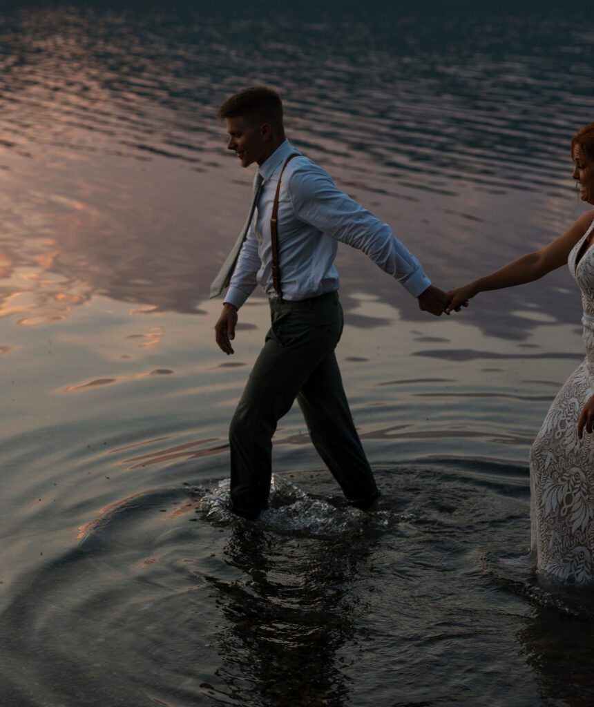 Bride and groom holding hands, walking in the water at sunset during their Glacier National Park elopement.