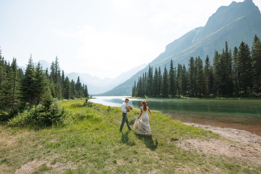 Bride and groom walking by the lake, surrounded by mountains and trees, during their Glacier National Park elopement.