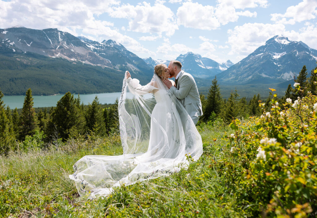 Bride standing on a mountain overlook, her veil flowing in the wind with stunning Glacier National Park scenery in the background.