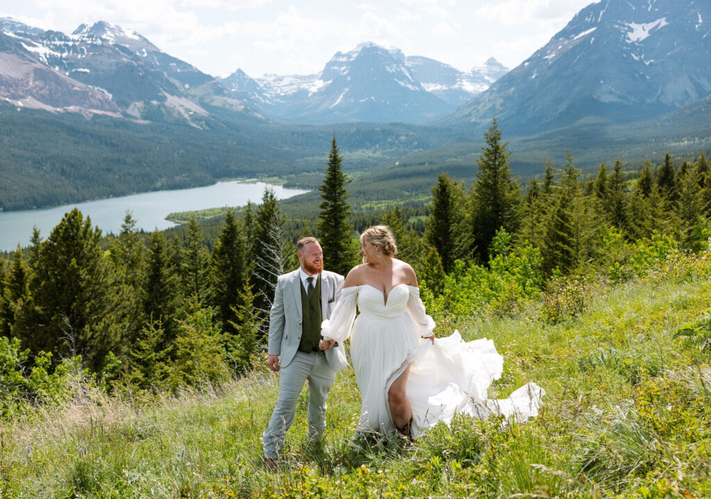 Bride and groom holding hands, walking through a field with mountains in the distance.