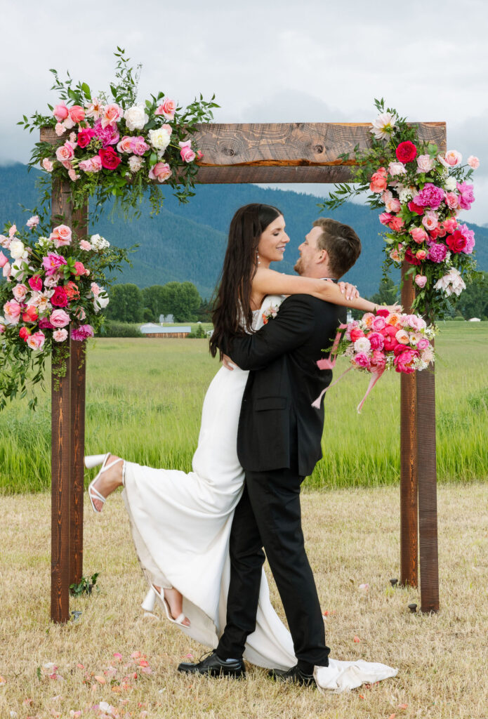 Bride and groom hugging under a floral arch with mountains behind them, captured by Montana wedding photographer Haley J Photo.