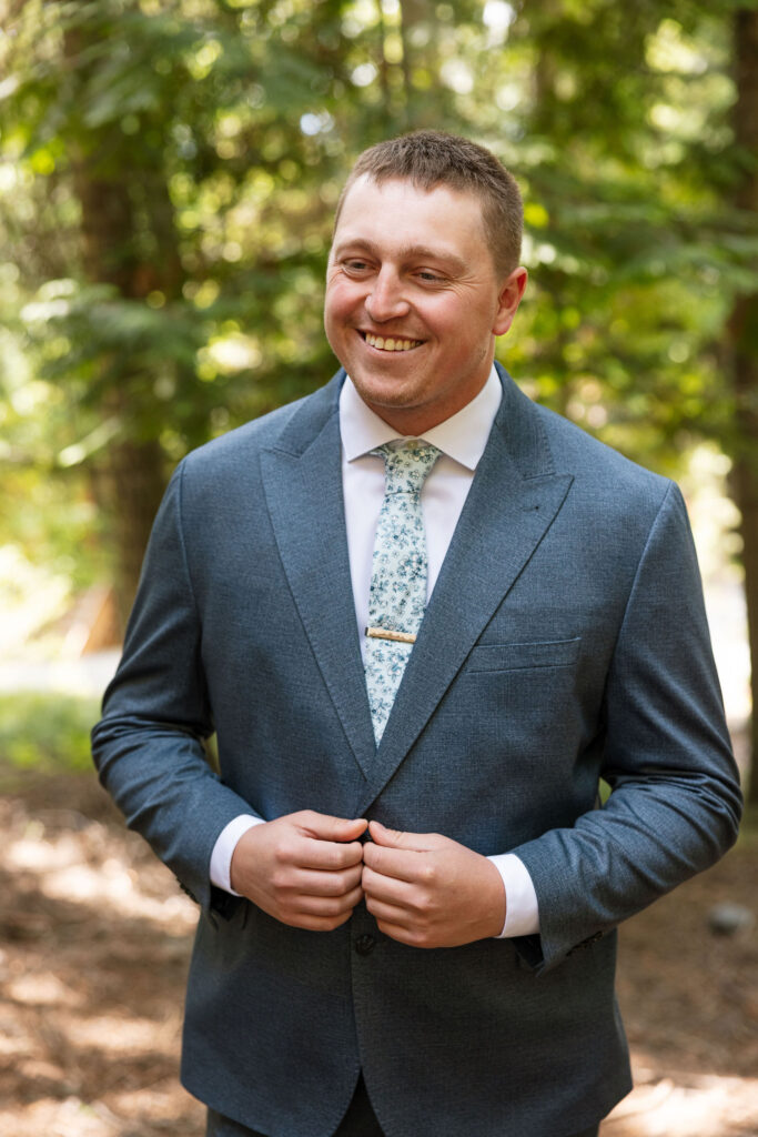 Groom smiling in a gray suit and floral tie, standing in the forest before the elopement ceremony.