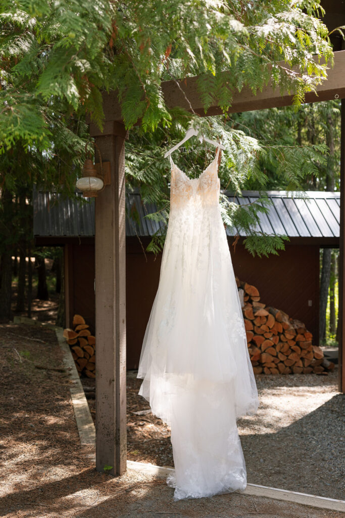 Wedding dress with lace detail hanging from a wooden beam outdoors, stacked firewood in the background.