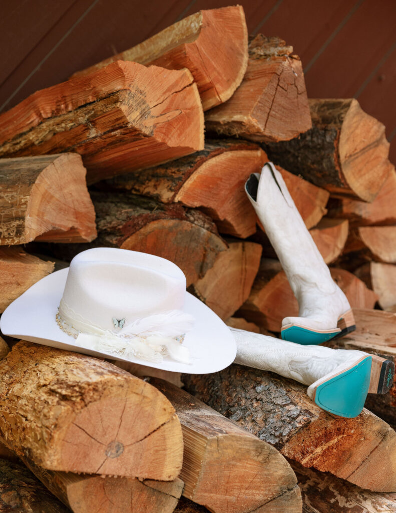 Bride’s white cowboy hat with feather accent and white leather boots with turquoise soles resting on firewood.