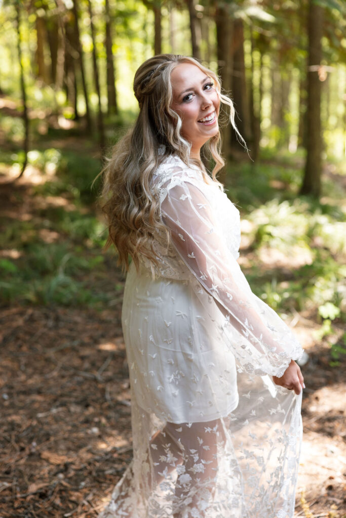 Bride smiling over her shoulder in a sheer floral robe, standing in a sunlit forest.