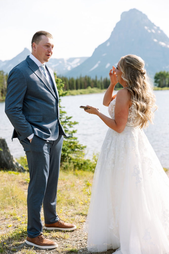 Bride wiping a tear while holding vow book, standing with groom in a gray suit at their Glacier National Park elopement.