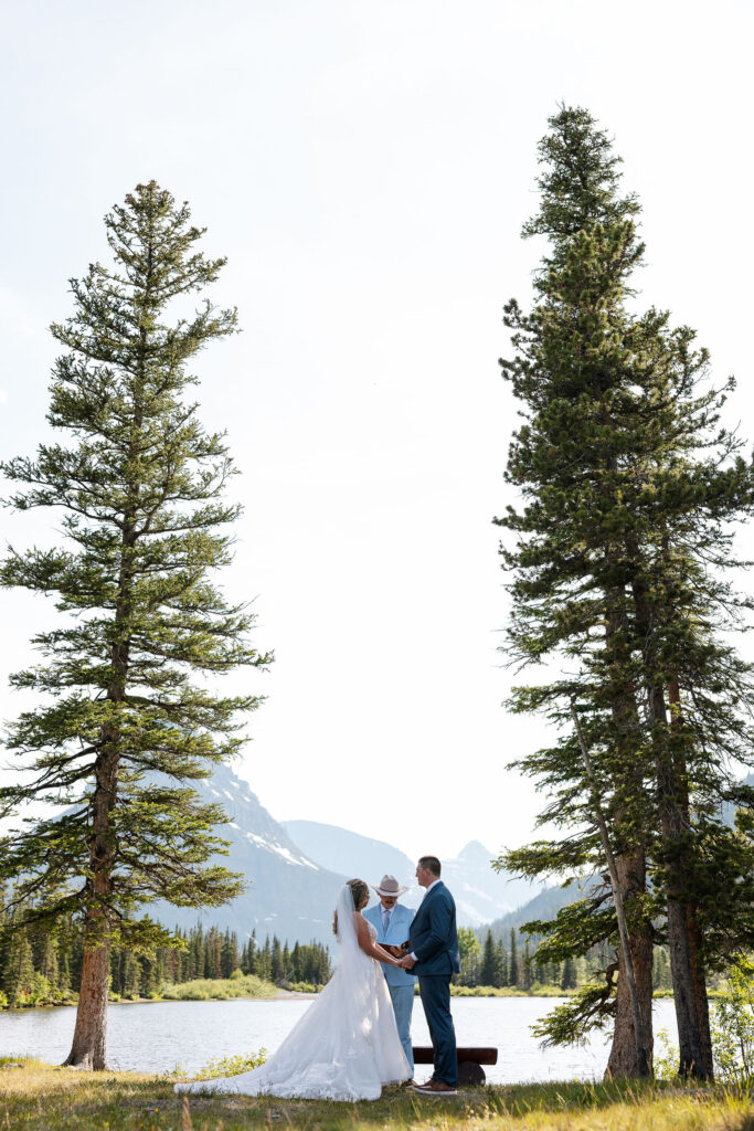 Bride and groom holding hands during their Glacier National Park elopement ceremony, standing between tall pine trees with mountain views.
