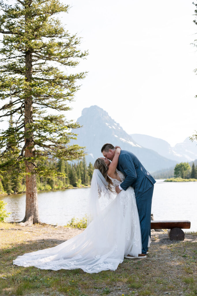Groom in gray suit dipping bride in lace gown for a kiss beside a mountain lake during their Glacier National Park elopement.