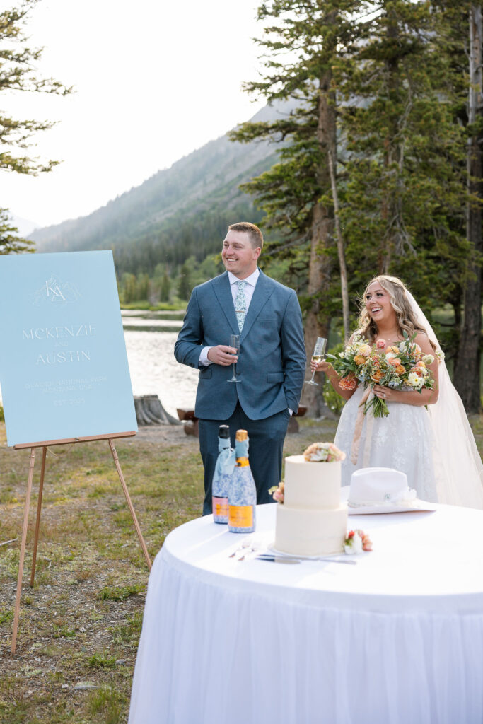 Bride and groom giving a toast beside their wedding cake and welcome sign at a lakeside Glacier National Park elopement.