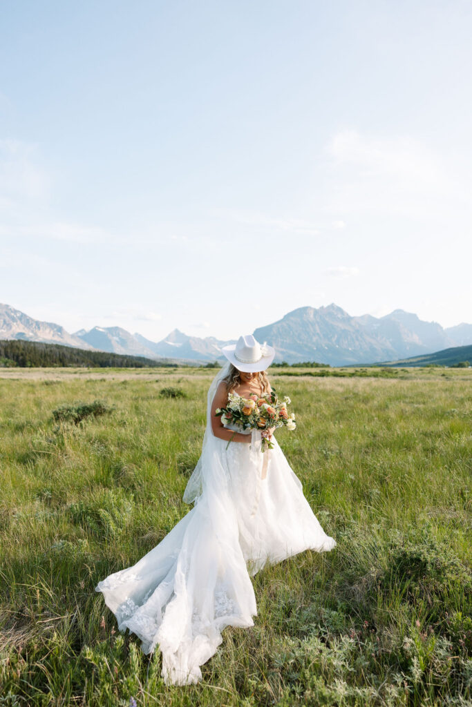 Bride in lace gown and cowboy hat holding a bouquet of peach and blush flowers, walking through an open field with mountain views in Glacier National Park.