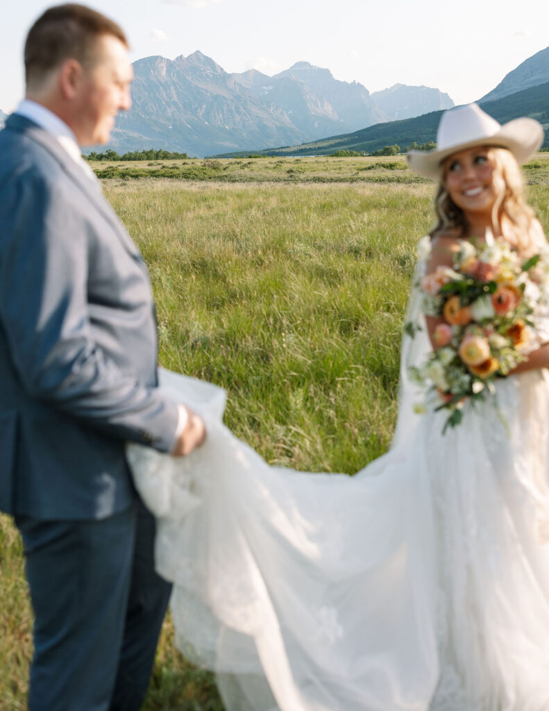 Groom holding the bride’s train as she smiles in a cowboy hat and lace gown during their Glacier National Park elopement.