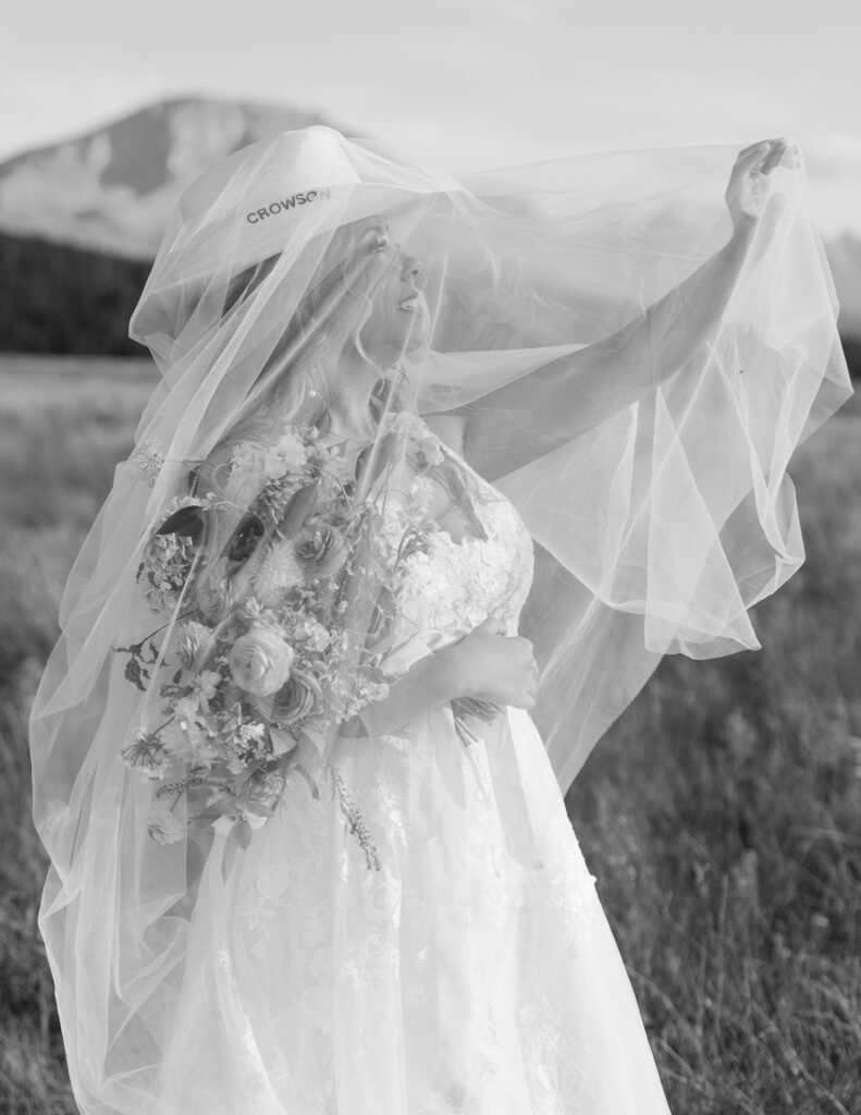 Black and white photo of bride lifting her veil while holding a bouquet of blush and peach flowers at a Glacier National Park elopement.