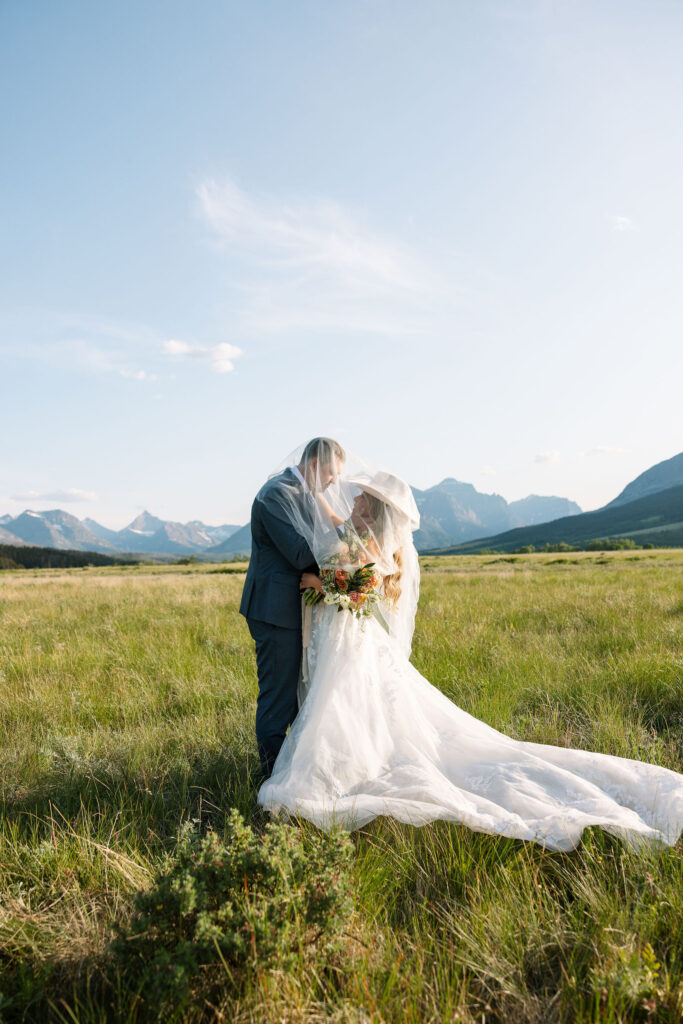 Bride and groom embracing in a wide open field with mountains in the distance at their Glacier National Park elopement.