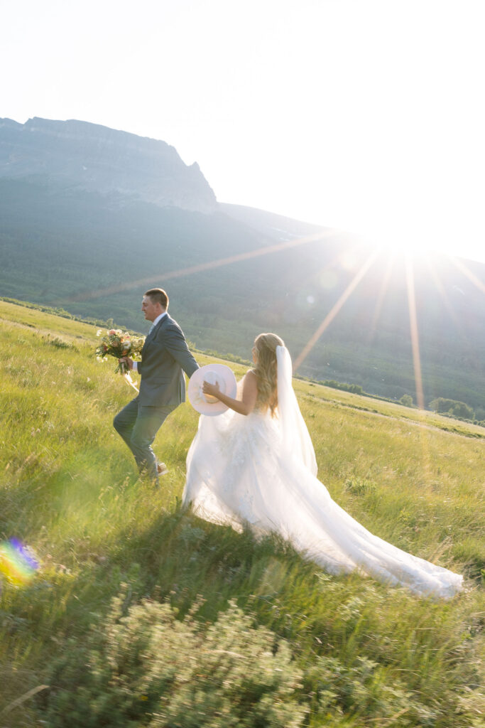 Couple walking hand in hand through a sunlit field at their Glacier National Park elopement, with the bride holding her bouquet and hat.