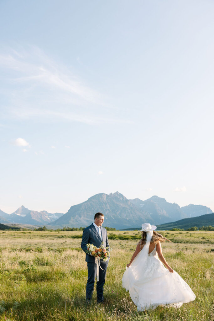 Bride in white cowboy hat twirling her gown in a meadow while groom holds bouquet, with mountain views in Glacier National Park.