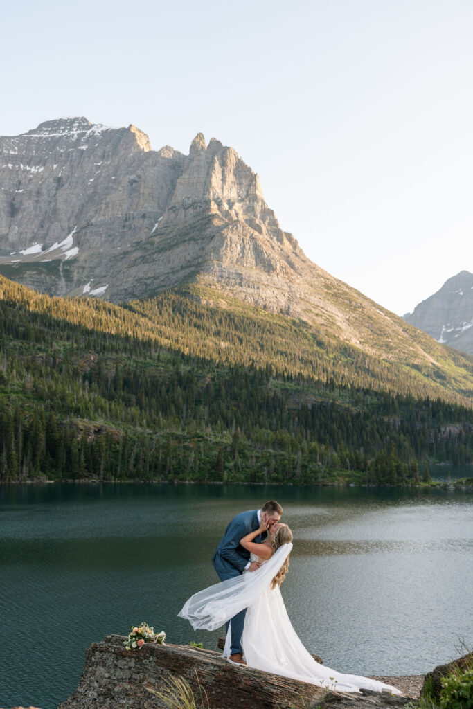 Bride and groom kissing on a cliff above a mountain lake during their Glacier National Park elopement.