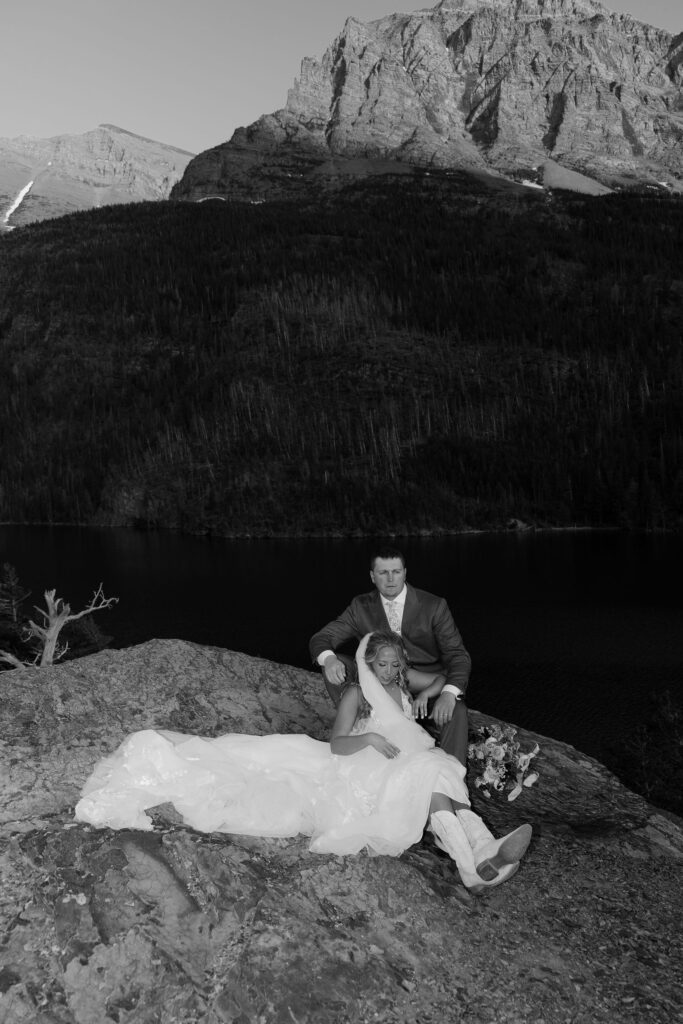 Black and white photo of bride and groom sitting together on a rock ledge overlooking a mountain lake at their Glacier National Park elopement.