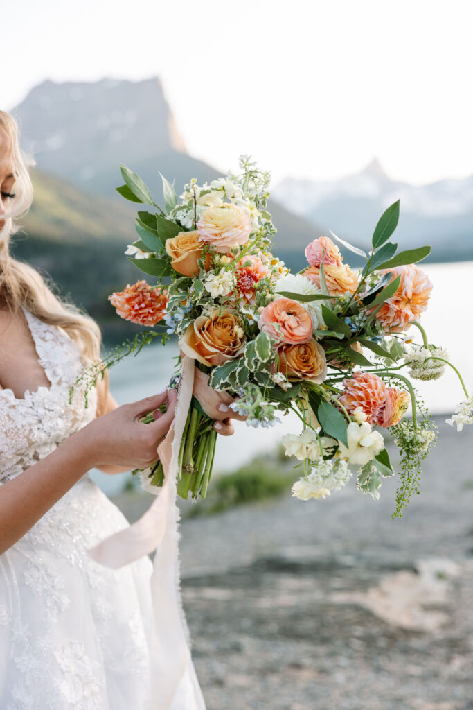 Close-up of bride holding bouquet with peach, orange, and cream flowers against a lake backdrop in Glacier National Park.