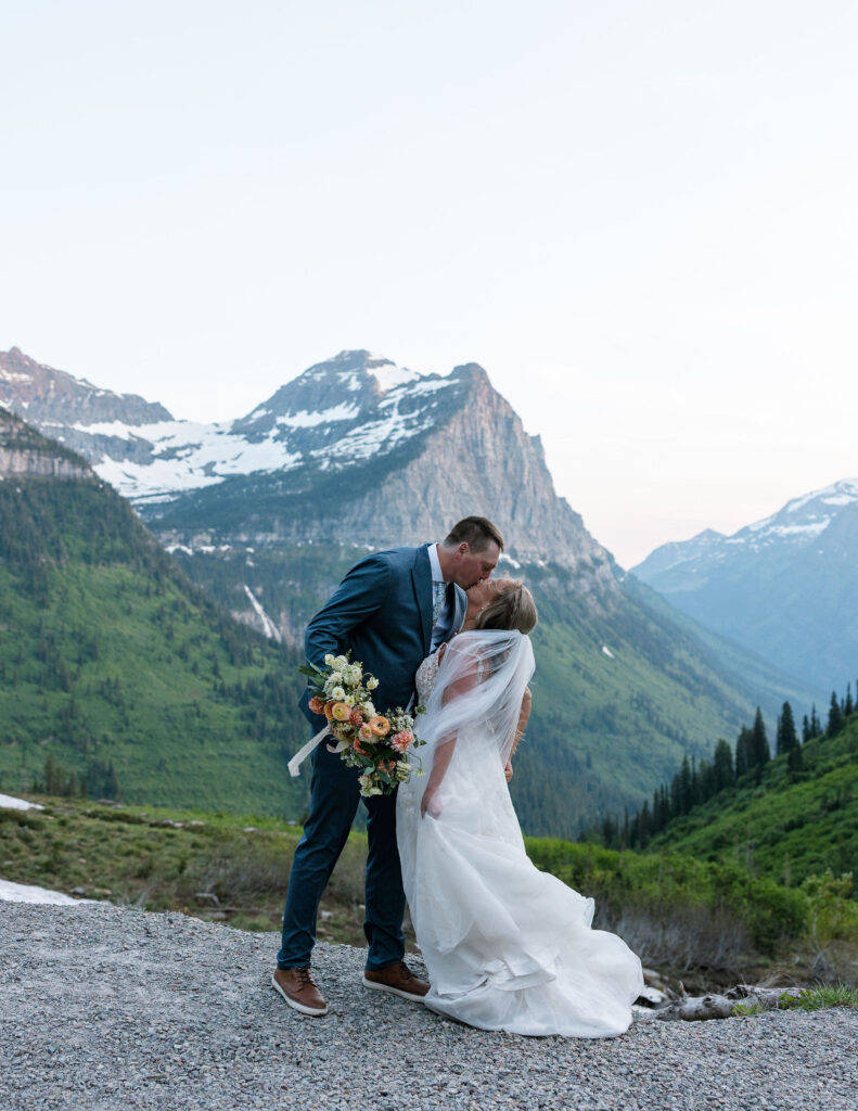 Bride and groom kissing on a mountain overlook with snowy peaks behind them during their Glacier National Park elopement.