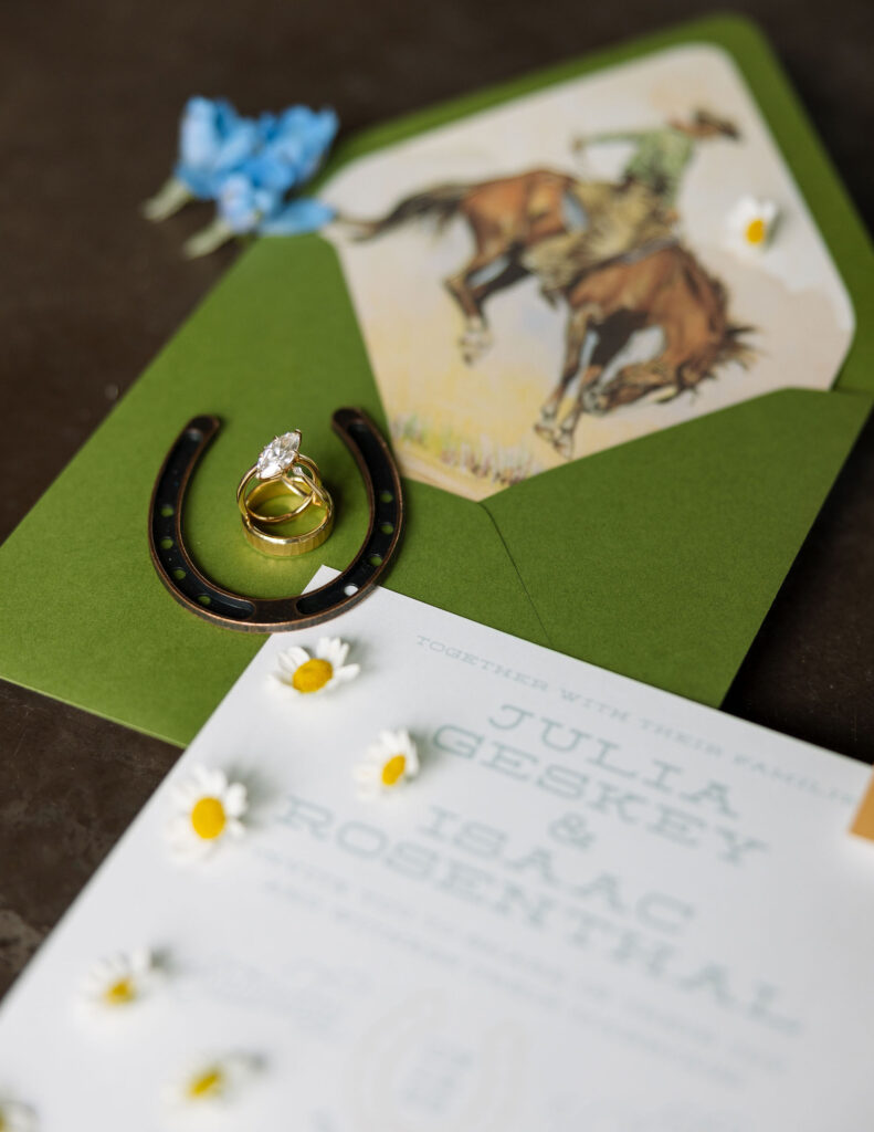 Western-inspired wedding flatlay with green envelope, illustrated cowboy invitation, horseshoe, and engagement rings, styled details from a Whitefish, Montana wedding.

