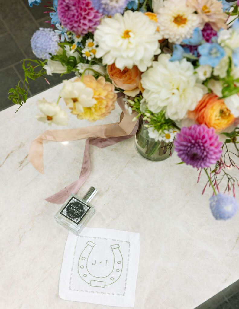 Colorful summer bridal bouquet with dahlias, ribbon-tied perfume, and custom embroidered horseshoe cocktail napkin — joyful wedding details in Kalispell, Montana.
