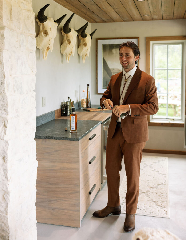 Groom in a rust-colored suit smiles while getting ready in a rustic Montana cabin, with western decor and whiskey on the counter. Pre-ceremony wedding morning.