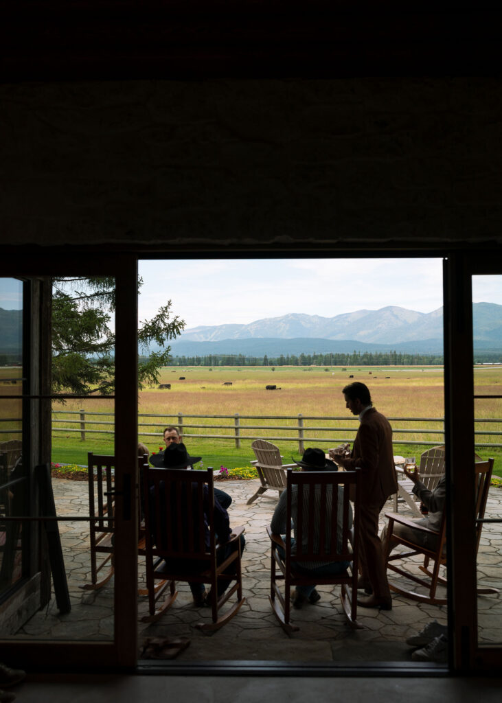 Groom and groomsmen gather on a ranch porch overlooking the mountains and grazing bison in Whitefish, Montana. Candid wedding morning moment filled with calm and connection.