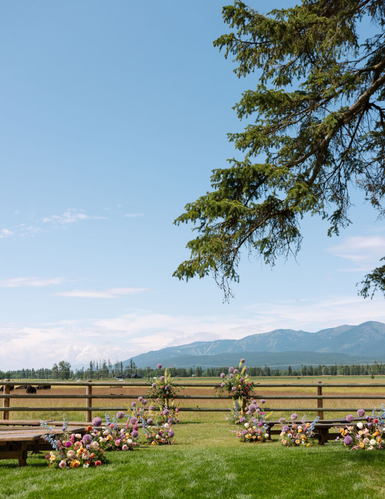 Outdoor ceremony setup on a ranch in Whitefish, Montana with vibrant floral arrangements, wooden benches, and mountain views under a clear summer sky.