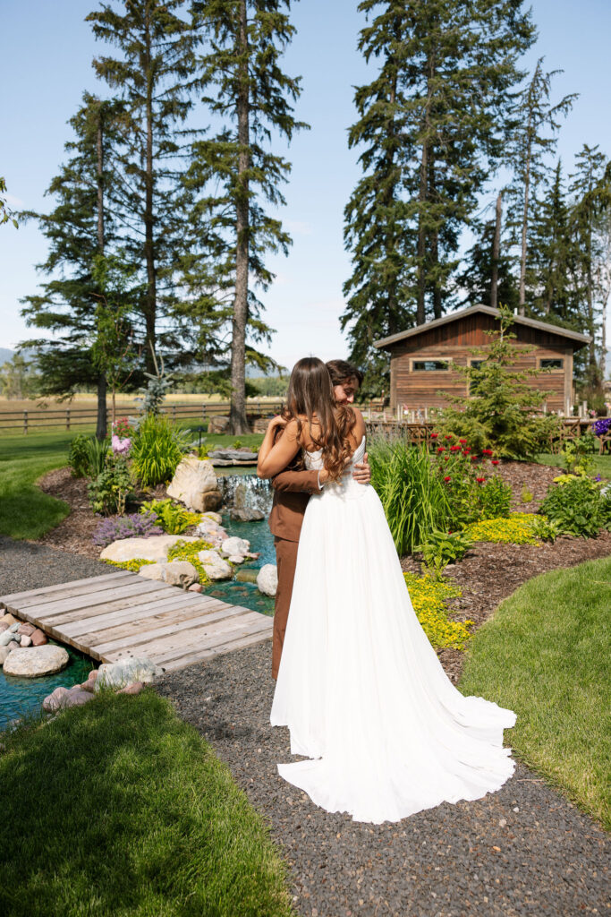 Bride and groom embrace during their emotional first look on a wooden bridge in a lush garden setting near Whitefish, Montana, with mountains in the distance. Joyful wedding photos.