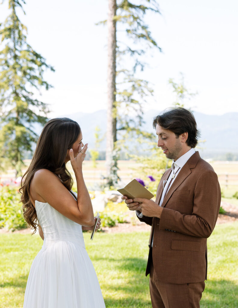 Bride and groom exchange emotional personal vows under the trees at a ranch wedding in Whitefish, Montana, with the bride wiping away a tear.