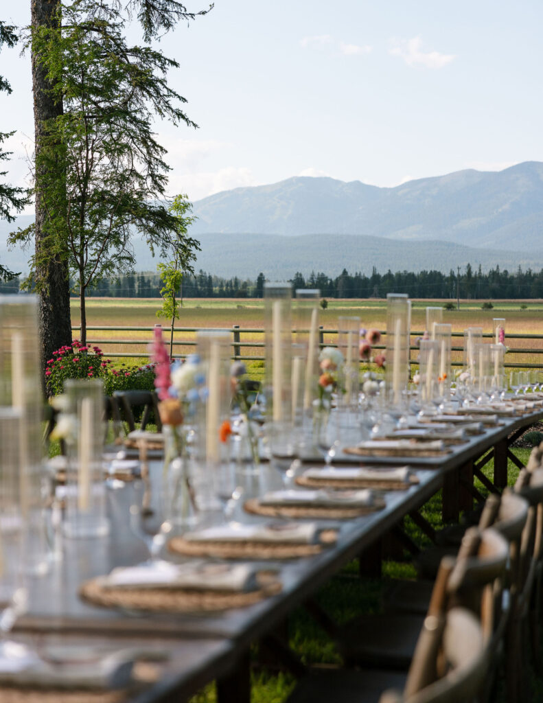 Rustic outdoor reception table set with glassware, florals, and woven chargers, overlooking Montana’s mountains and open ranch land.