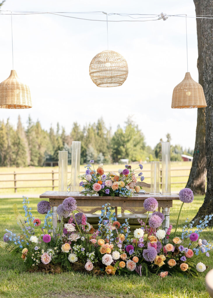 Rustic sweetheart table adorned with colorful florals, glass pillar candles, and woven pendant lights at an outdoor Montana wedding reception.