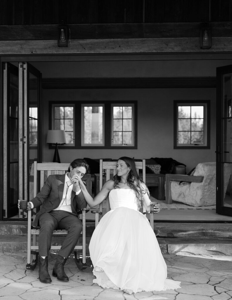 Bride and groom share a quiet moment on a porch after their ceremony, with the groom gently kissing the bride’s hand — a candid black-and-white photo capturing deep connection.