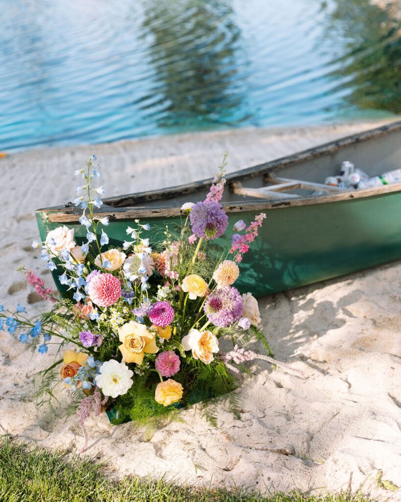 Wildflower floral arrangement on the sand beside a green canoe, set against a clear blue lake. Whimsical wedding detail at a lakeside venue in Montana.