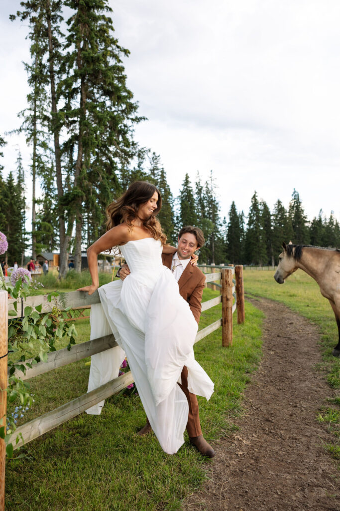 Groom lifts the bride over a wooden fence at a ranch wedding in Montana, both laughing as horses look on in the background. A candid, adventurous moment.