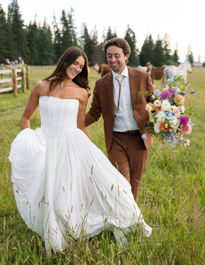 Bride and groom walk hand-in-hand through a pasture, smiling with wildflowers and horses behind them. Joyful newlywed portraits at a Montana ranch wedding.