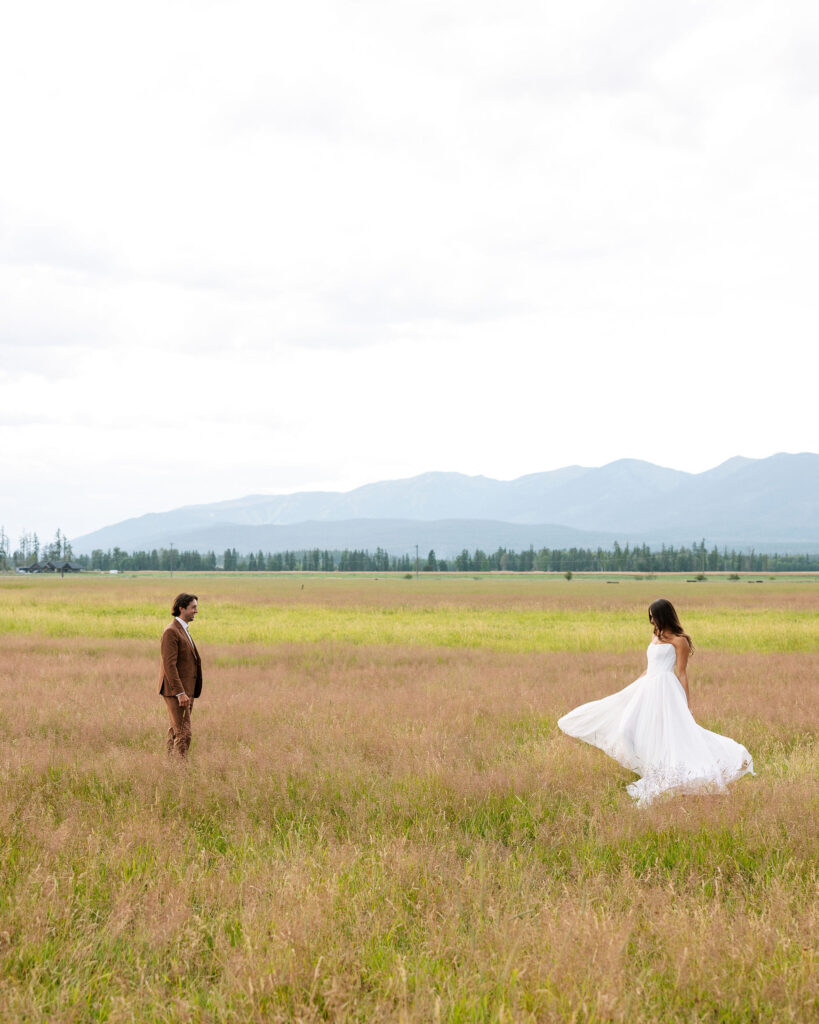 Bride twirls in a wide open Montana field with mountains in the distance, while the groom watches with a smile. Dreamy and joyful wedding portrait.