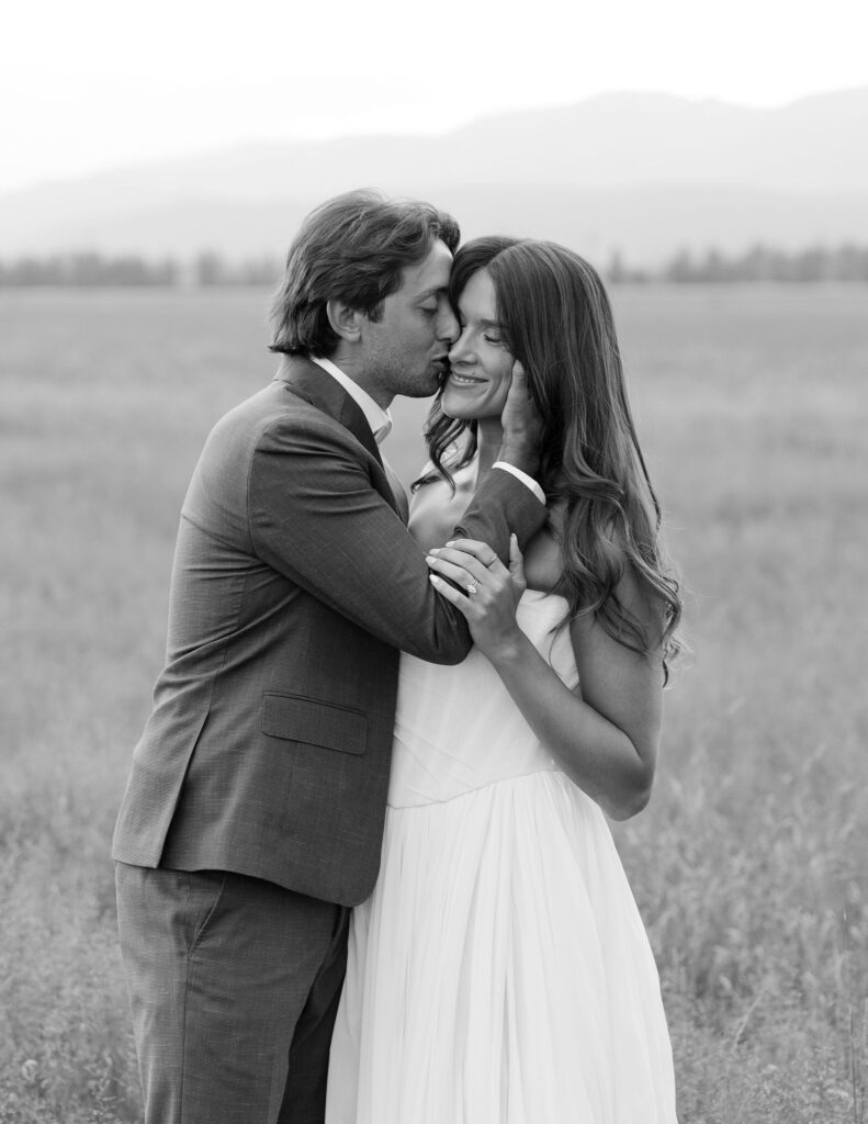 Groom kisses the bride’s forehead during an intimate moment in a quiet Montana meadow. A timeless black-and-white portrait full of love and tenderness.
