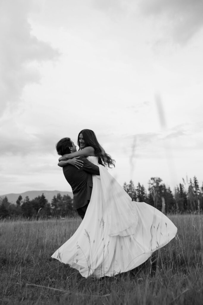 Groom lifts bride in an open Montana field, both laughing, as her dress flows in motion. A candid, joyful moment captured at sunset.

